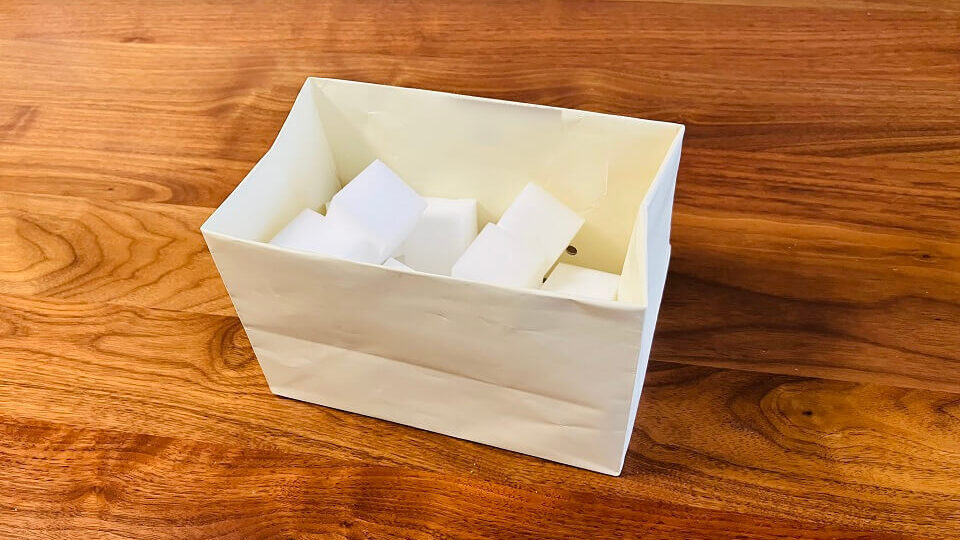 クリーム色の紙袋で作った収納ケースに、日用品（メラミンスポンジ）が入っている。